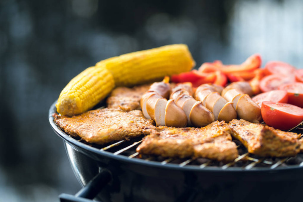 Close up van voedsel op een houtskool waterkoker grill grillen. Worst, vlees, maïs en groenten in openlucht barbecue in de zomer. BBQ-partij, achtertuin cookout of camping concept. - Foto, afbeelding