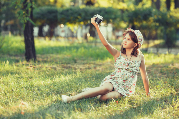 niña es interpretado por la cámara de fotos sentado en la hierba en el parque. Haciendo Selfie y fotografiando el mundo alrededor
 - Foto, Imagen