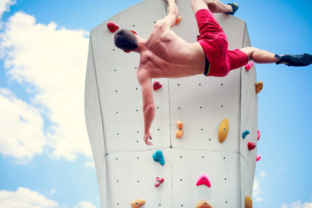 Photo from back of young athletic guy exercising on wall for climbing against blue sky with clouds - Foto, Imagen