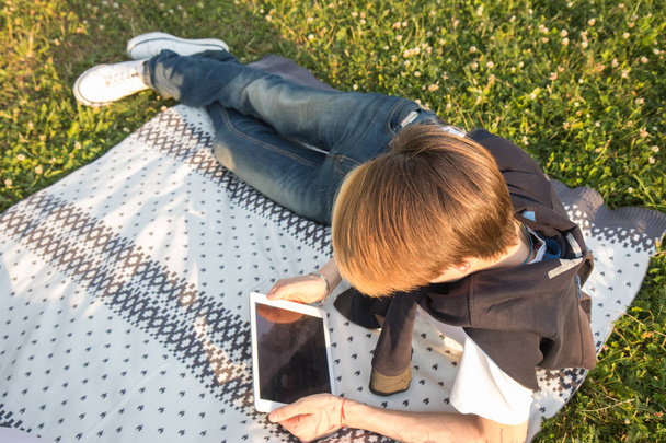Young guy with tablet  sitting  in the park  on the rug  green grass in park summer day - Valokuva, kuva