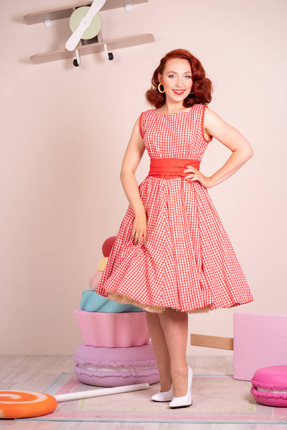Beautiful red-haired pinup smiling happily girl posing in a retro red dress and white high heels - Foto, Imagem