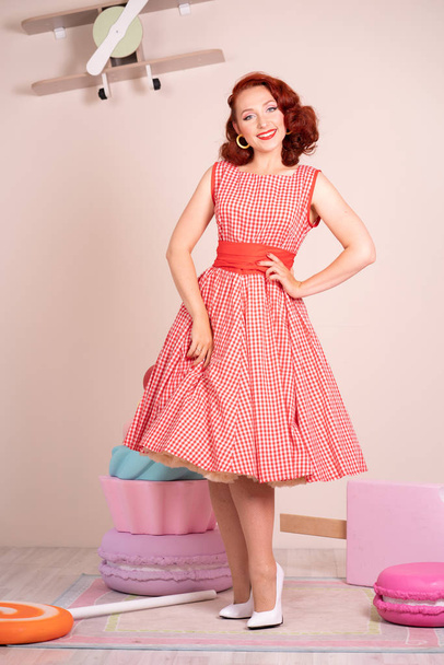 Beautiful red-haired pinup smiling happily girl posing in a retro red dress and white high heels - Foto, Bild
