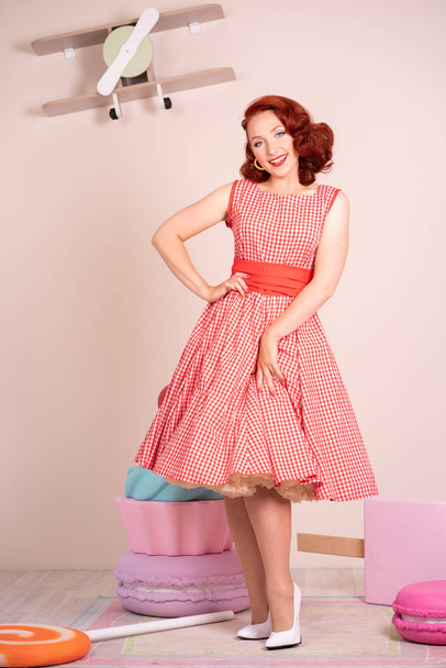 Beautiful red-haired pinup smiling happily girl posing in a retro red dress and white high heels - Photo, Image