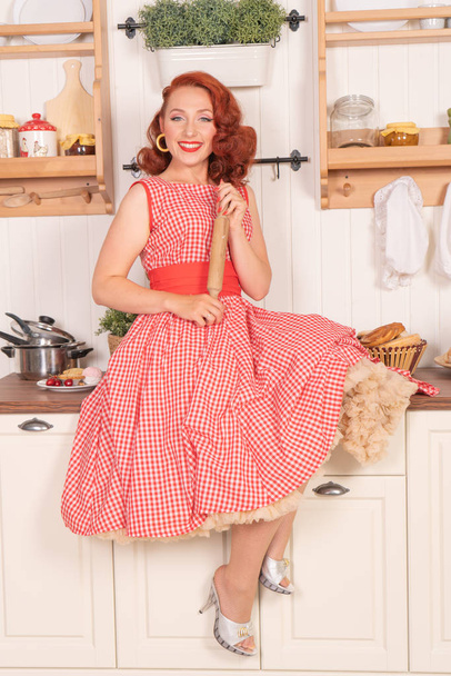 beautiful red-haired pinup smiling happily girl posing in a retro red dress in the kitchen alone - Foto, imagen