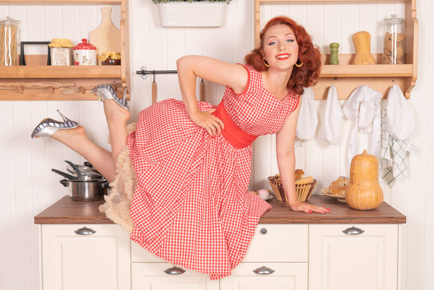 beautiful red-haired pinup smiling happily girl posing in a retro red dress in the kitchen alone - Photo, Image