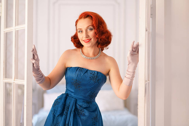eine luxuriöse Pin-up-Dame in einem blauen Vintage-Kleid steht in der Tür ihres Schlafzimmers und lädt Sie ein, ins Haus zu kommen - Foto, Bild