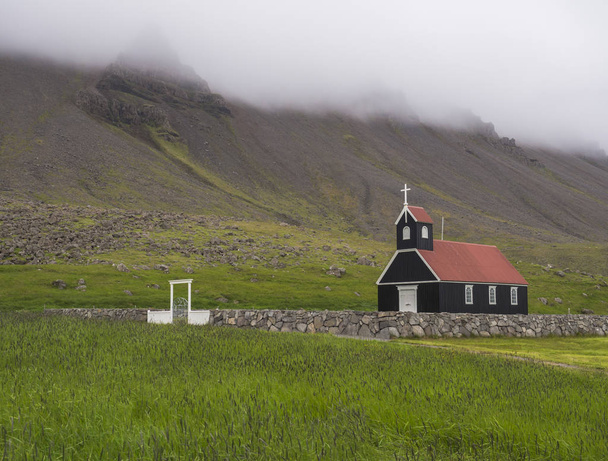 Saurbaejarkirkja black wooden red roof church on green grass field, steep hills in fog, Raudisandur, iceland west fjords - Zdjęcie, obraz