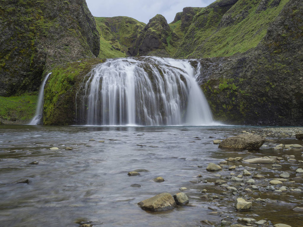 Bela cachoeira Stjornarfoss no sul da Islândia, perto do acampamento Kirkjubeajarklaustur, com rochas, penhascos e colinas verdes, musgo e céu azul, borrão de movimento de longa exposição
 - Foto, Imagem