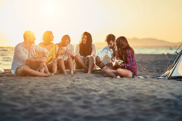 Group of young friends with guitar at beach. friends relaxing on sand at beach with guitar and singing on summer sunset - Photo, Image