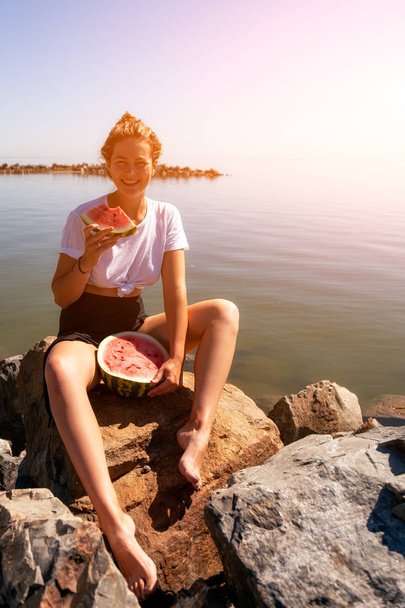 joven hermosa mujer riendo alegremente, sonriendo, sosteniendo entre las piernas una gran sandía y comiendo, sentado rocas de piedra en un día de verano cerca del mar
 - Foto, imagen