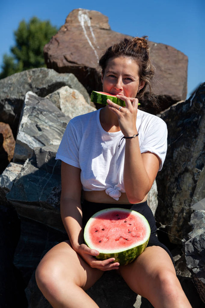 fiatal gyönyörű nő nevetve, boldogan, mosolyogva, gazdaság, a lába között egy nagy görögdinnye és evés, kő sziklák ül egy nyári nap, a tenger közelében - Fotó, kép