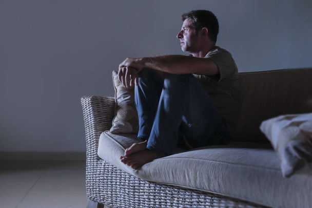 lifestyle dramatic light portrait of young sad and depressed man sitting at shady home couch in pain and depression feeling lost looking away thoughtful and pensive suffering anxiety problem - Фото, изображение