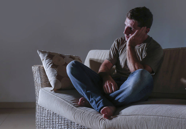lifestyle dramatic light portrait of young sad and depressed man sitting at shady home couch in pain and depression feeling lost looking away thoughtful and pensive suffering anxiety problem - Фото, зображення