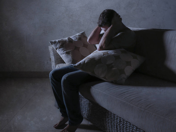 lifestyle dramatic light portrait of young sad and depressed man sitting at shady home couch in pain and depression feeling stressed and desperate crying alone suffering anxiety crisis - Fotoğraf, Görsel