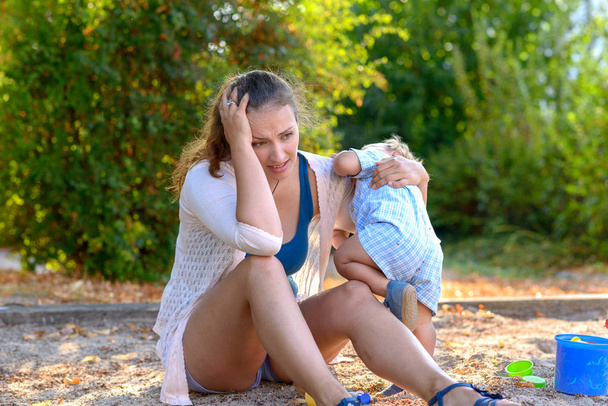 Jeune mère stressée avec un bébé garçon difficile regardant le sol dans une aire de jeux alors que l'enfant agit alors qu'elle le tient près d'elle
 - Photo, image