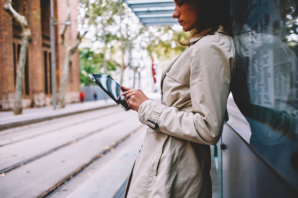 Cropped side view of young woman in trendy coat checking mail and reading notification on touch pad using free 4G internet.Pensive female blogger updating profile on tablet standing on tram stop - Φωτογραφία, εικόνα