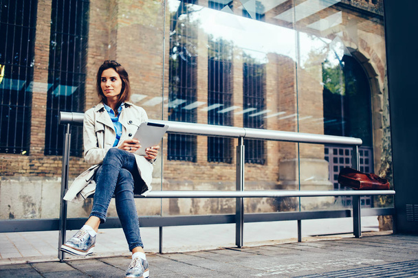 Thoughtful stylish hipster girl installing application on modern touch pad via free 4G internet connection while looking on side waiting on tram.Pondering young woman with tablet sitting on bus stop - Foto, Imagen