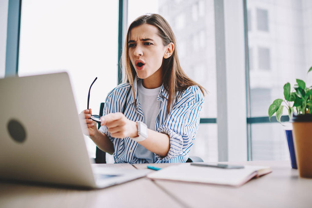 Emotional woman stressed while watching video on laptop computer sitting in office interior, shocked female pointing on netbook irritated about application failure during working process at deskto - 写真・画像