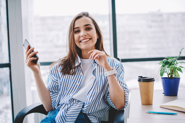 Cheerful caucasian woman satisfied with good wireless connection in office holding smartphone for chatting , happy female employee looking at camera using mobile phone during rest on coffee brea - Фото, изображение