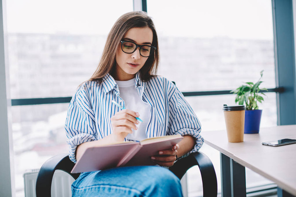 Clever female employee in eyewear writing in notepad concentrated on working process in office, serious caucasian smart woman making notes of ideas for article sitting at comfortable working plac - Foto, immagini