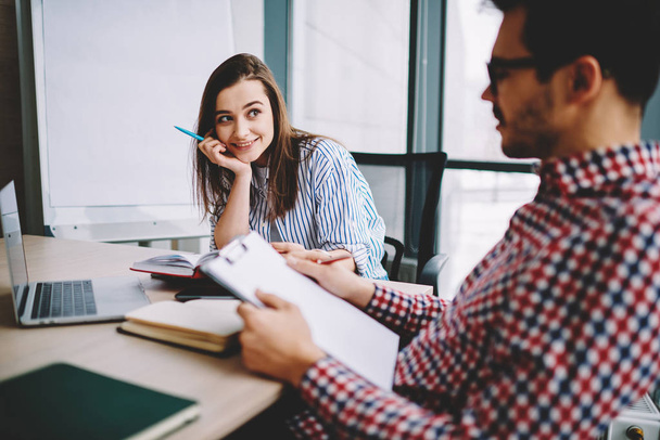 Cheerful woman looking at male colleague reading report and analyzing information during cooperation on startup in office,smiling female employee satisfied with working process with man coworke - Foto, Bild