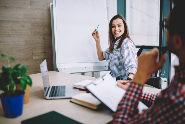 Smiling young woman pointing on flip chart explaining ideas to her male colleague cooperating on making strategy for startup, cropped image of cheerful female designer using board consulting with bos - Zdjęcie, obraz