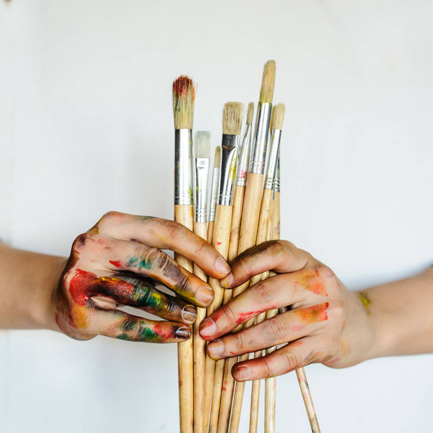 Primer plano de las manos de un artista / pintor sosteniendo pinceles. Sucio pintado con pintura de colores sobre fondo blanco
 - Foto, imagen