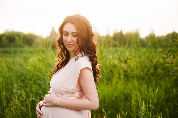Feliz mujer embarazada caminando en el campo verde en el parque al amanecer con una cálida luz de fondo
 - Foto, Imagen