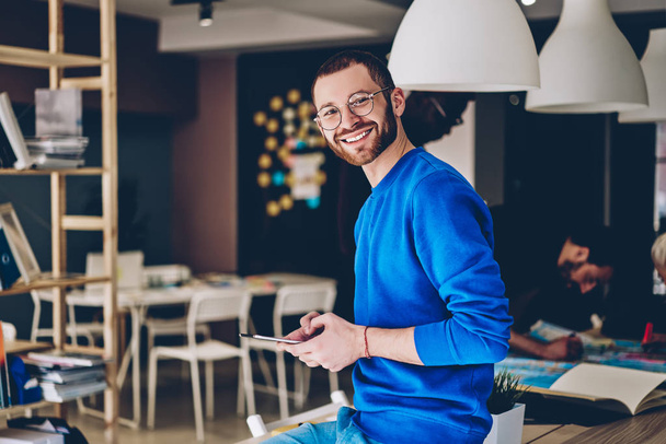 Portrait of cheerful young man in casual wear smiling at camera while updating software on modern touch pad device using wireless 4G internet.Positive hipster guy installing app on tablet in office - Foto, Bild