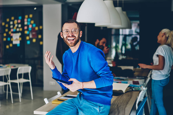 Positive caucasian young man laughing while updating profile on modern touch pad device using wireless 4G internet in office.Emotional cheerful hipster guy having fun while holding digital tablet - Foto, Bild
