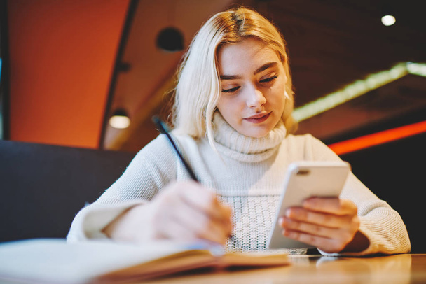 Young woman making notes using organizing application on cellular for planning tasks and learning process, female writing in notepad information from web page browsed on smartphone in cafe interior - Foto, Imagem