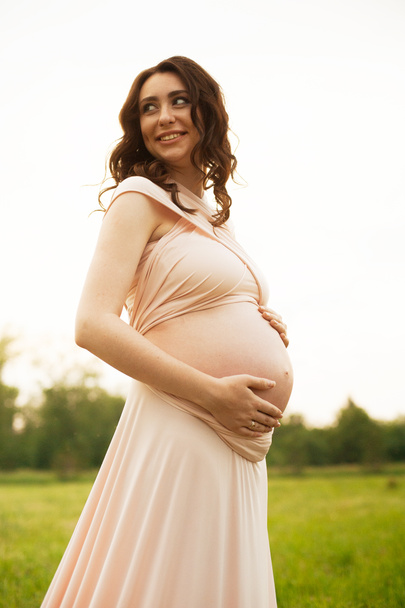 Счастливая беременная женщина гуляет по зеленому полю в парке на рассвете с теплым задним светом на заднем плане
. - Фото, изображение
