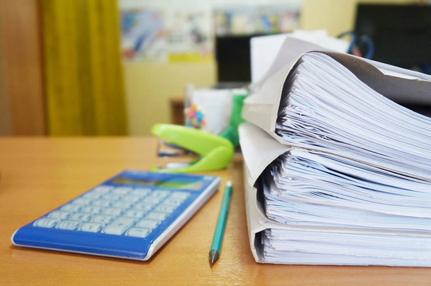 Бумаги на столе. Куча документов в картонных папках, калькулятор и канцелярские принадлежности на столе, вид сбоку
            - Фото, изображение