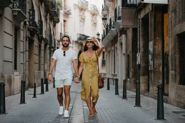 Pretty brunette girl with her boyfriend with beard walking holding hands on the old small European street in Spain on the sunset - Foto, afbeelding