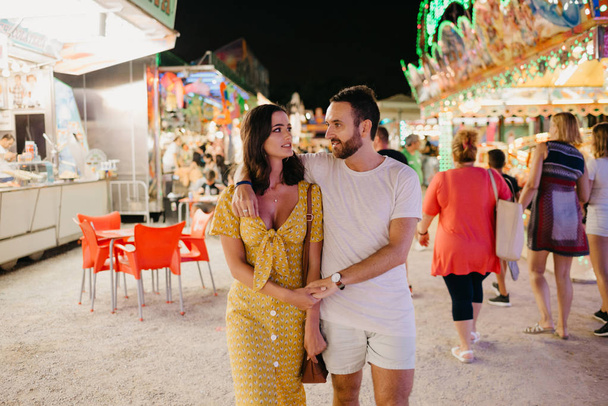 Симпатична брюнетка дівчина зі своїм хлопцем з бородою ходьба холдингу руки на ярмарку в Іспанії у вечірній час - Фото, зображення