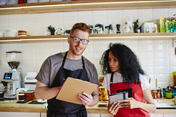 Caucasian male waiter and african american waitress checking order of client and looking at camera standing in own modern cafe interior.Two friends entrepreneurs making plan of cost of own business - Zdjęcie, obraz