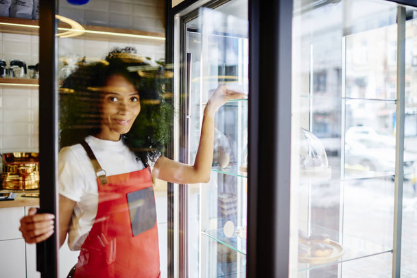 Portrait of positive african american young woman working professional confectioner in own coffee shop and taking dessert from refrigerator with sweet cakes for sell during working process - Fotó, kép
