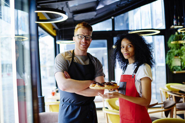 Portrait of positive caucasian barista with crossed hands teamworking with cheerful african american female confectioner which holding plate with tasty delicious pastries standing in common bakery - Foto, Bild
