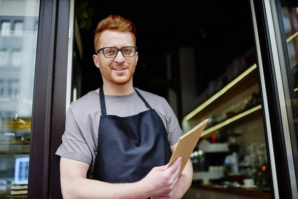 Portrait of cheerful caucasian waiter in apron holding menu in hands and inviting to own cafeteria.Positive handsome male owner of coffee shop standing outdoors and showing hospitality - Foto, imagen
