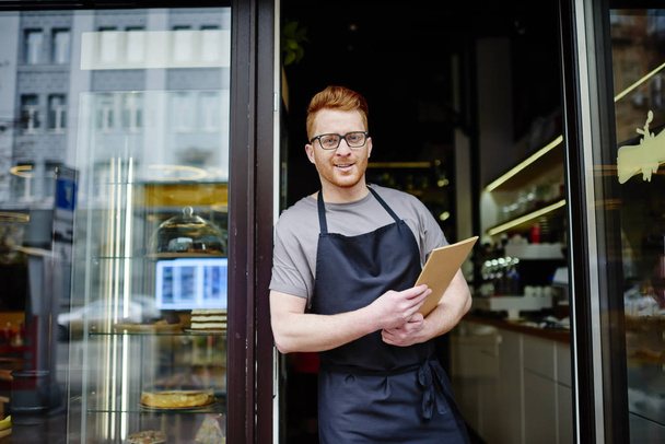 Portrait of cheerful owner of bakery holding menu and inviting to shop.Positive waiter in apron smiling at camera while standing outdoors at entrance to cafe and showing hospitality - Foto, imagen