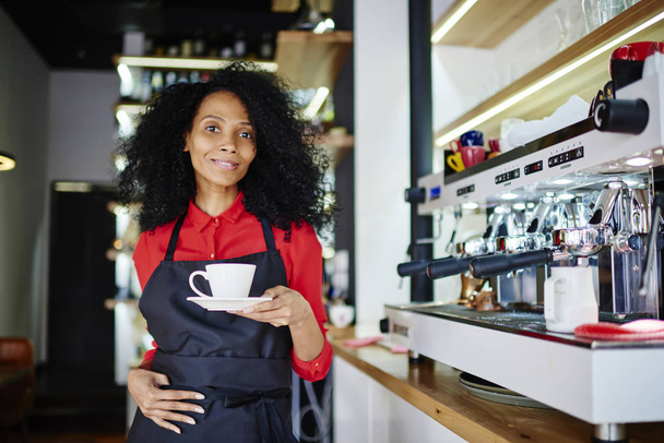 Half length portrait of african american young woman working barista and holding cup of aroma caffeine beverage in hands while smiling at camera standing near modern coffee machine in cafe - Fotoğraf, Görsel