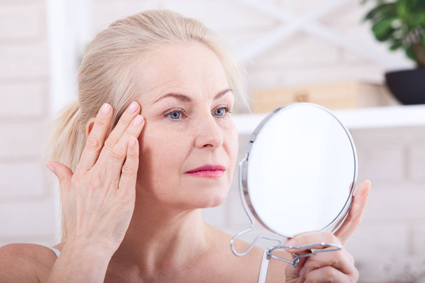 Forty years old woman looking at wrinkles in mirror. Plastic surgery and collagen injections. Makeup. Macro face. Selective focus - Foto, Imagen