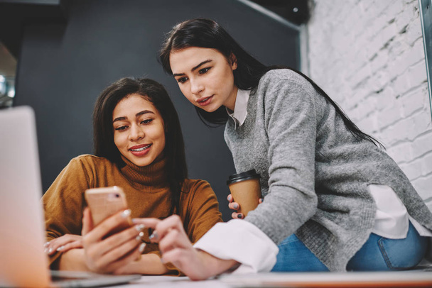 Hipster girl helping her friend installing useful app for learning languages during studying in college, colleagues booking tickets via smartphone online on coffee break together choosing best price - Photo, Image