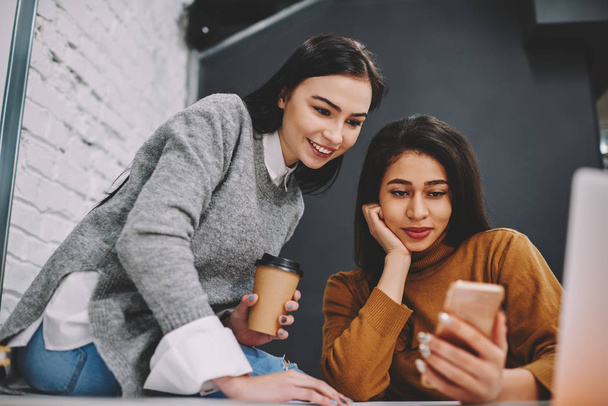 Positive hipster girls viewing photos in social networks profile using smartphone and wifi connection, smiling female best friends watching video on cellular during coffee break in college campu - Photo, Image