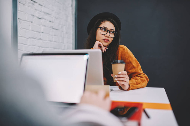 Bild von der Studentin, die am Laptop arbeitet, während ihr kontemplativer Freund mit coolem Hut über erfolgreich bestandene Prüfungen träumt, junge attraktive Frau in der Kaffeepause grübelnd - Foto, Bild