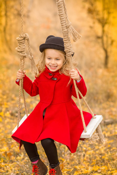 little girl in a red coat swings on a swing and makes funny faces - Фото, изображение