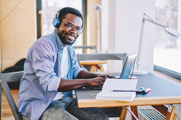 Cheerful dark skinned male student listening audio book while learning using technology in campus, prosperous guy enjoying playlist songs in stereo equipment during freelance job on laptop compute - Zdjęcie, obraz