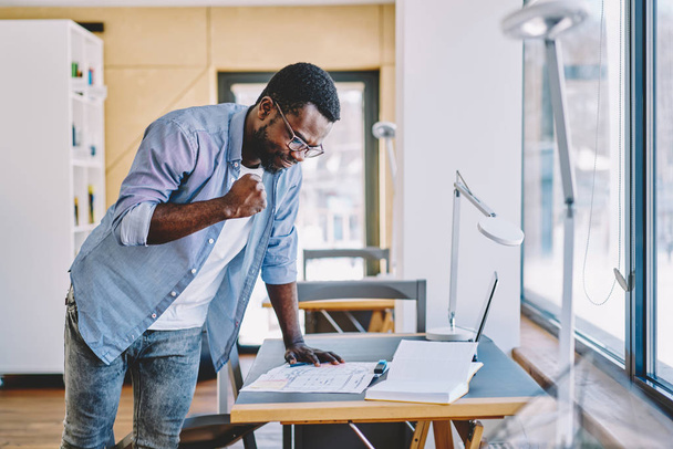 Serious african american hipster guy satisfied with completed coursework standing at desktop, skilled dark skinned male architect looking at accomplished creative sketch for project in coworking spac - 写真・画像
