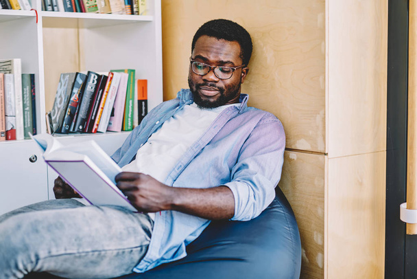 Serious clever african american student in spectacles for vision correction reading interesting book on leisure thinking about plot,dark skinned hipster guy analyzing information from literature - Foto, imagen