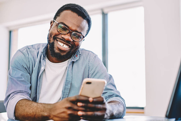 Cheerful african american hipster guy laughing at funny video on smartphone during free time, happy dark skinned man enjoying good connection of 4G internet for watching movie on mobile phon - Fotó, kép
